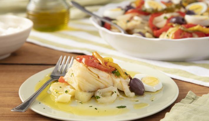 Imagem de um prato branco com uma porção de bacalhoada com ovos, pimentões, azeitona preta, tomate e cebola com um garfo ao lado. Ao fundo, uma travessa de bacalhoada e um vidro de azeite.