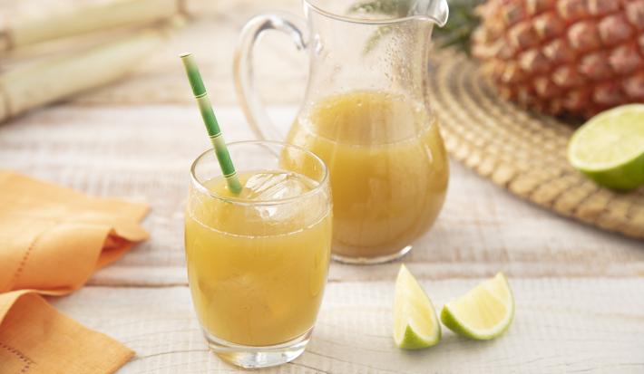 Fotografia em tons de laranja e verde em uma bancada de madeira clara, um pano laranja, um jogo americano de palha com um abacaxi e limão em cima dele. Ao centro, um copo de vidro com caldo de cana dentro e uma jarra com o restante do caldo de cana.