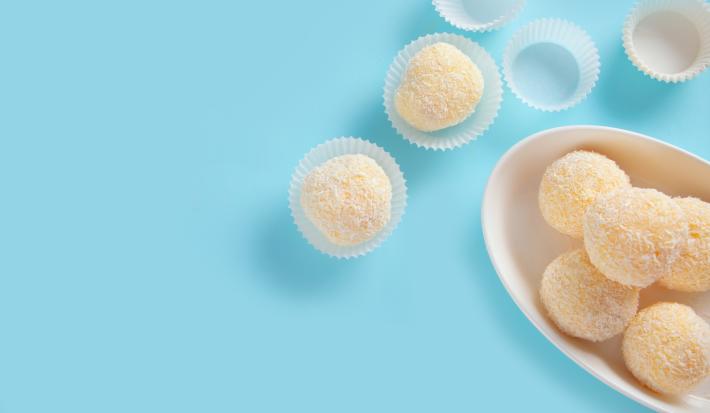 Fotografia vista de cima de cinco docinhos recheados com geleia de damasco e passados no coco dentro de um recipiente oval e fundo de cor branca. Ao lado tem quatro forminhas de brigadeiro, e duas estão com o docinho, e estão sobre uma mesa azul.