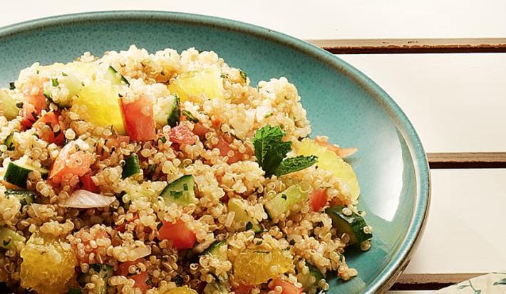 Fotografia em tons de verde e amarelo, com porcelana verde contendo tabule de quinoa com legumes e gomos de laranja, decorado com folhas de hortelã, tudo sobre bancada de madeira bege.