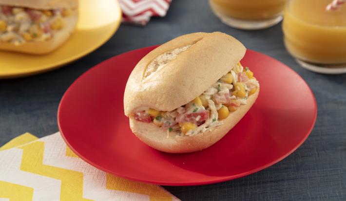 Foto da receita de sanduíche saudável sem glúten, montado em um pão francês recheado com um creme de frango, milho, sobre um prato vermelho e uma mesa com tecidos em tons amarelo e vermelho, ao lado de dois copos de suco e mais um sanduíche em outro prat
