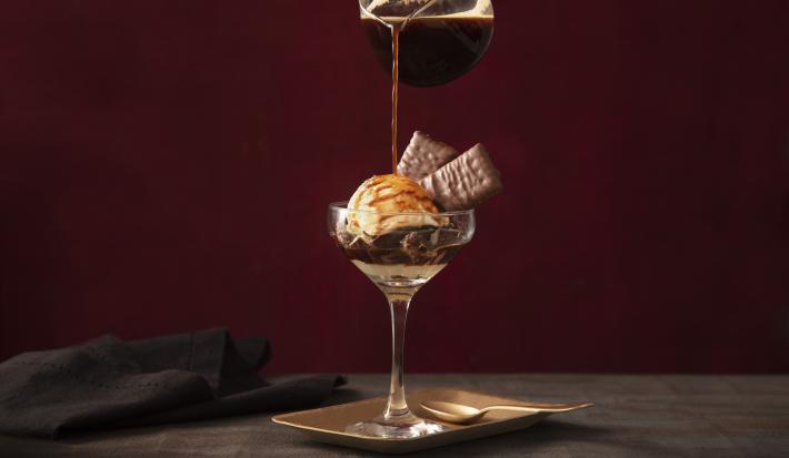 Fotografia em tons de preto de uma bancada de madeira com um prato quadrado, sobre ele uma taça com o affogato e uma jarra de café adicionando café á taça. Ao lado um paninho preto.
