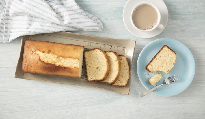 Fotografia em tons de azul em uma bancada de madeira clara com um prato redondo raso azul com uma fatia de bolo de Leite Moça. Ao lado, um recipiente retangular bege com o bolo inteiro.