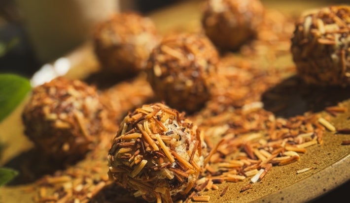 Fotografia em tons claros sob o sol de um prato grande, raso e de tom marrom claro. Sobre o prato tem alguns brigadeiros enrolados e passados no coco queimado, com mais coco queimado no prato. Ao fundo tem uma lata do leite ninho zero lactose.