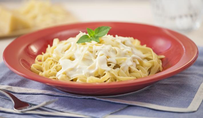 Fotografia em tons de vermelho e azul em uma bancada de madeira com jogos americanos azul e um prato vermelho com o macarrão ao molho branco dentro. Ao fundo, uma peça de queijo parmesão em fatias.