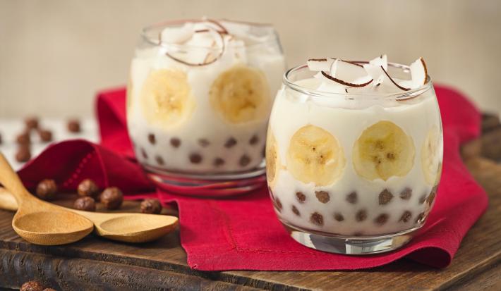 Fotografia em tons de vermelho e marrom em uma bancada branca com uma tábua de madeira escura, um pano vermelho, duas taças de vidro com o docinho de banana com Nescau Cereal.