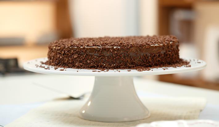 Imagem de um bolo redondo coberto com chocolate granulado sobre uma boleira branca.