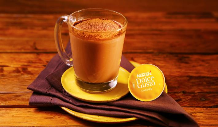 Fotografia em tons de amarelo em uma bancada de madeira escura, um prato amarelo redondo, um pano marrom e uma xícara de vidro com o chocafé com chocolate dentro dela.