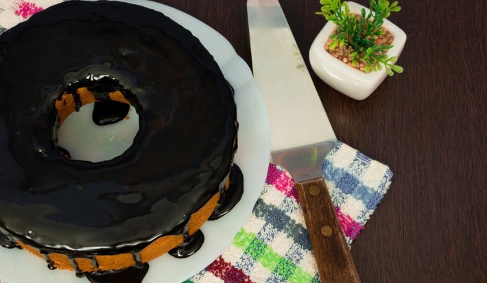 Um bolo de cenoura coberto com uma rica cobertura de chocolate, posicionado em um prato branco sobre uma superfície de madeira escura. Ao lado, um servidor de bolo de cabo de madeira e um pequeno vaso com uma planta artificial.