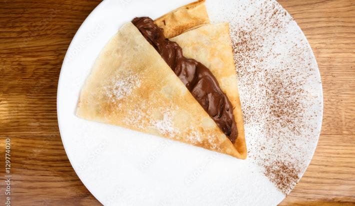 Fotografia vista de cima de uma panqueca com calda quente de mel e chocolate. A panqueca está em um prato branco raso com um pouco de cacau em pó peneirado por cima. O prato está sobre uma mesa de madeira.