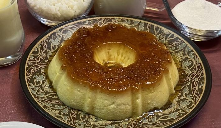 Foto em tons de bege escuro da receita de pudim de arroz inteira servida sobre um prato grande decorado. Ao fundo há alguns recipientes com arroz, açúcar, leite ninho e leite MOÇA.