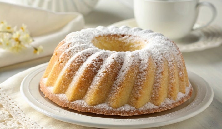Um bolo tipo bundt (em formato de anel) com uma casquinha dourada e cobertura de açúcar de confeiteiro. Está servido em um prato escuro ao lado de uma xícara de café, criando um clima acolhedor e caseiro.