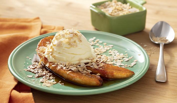 Fotografia em tons de branco em uma bancada de madeira de cor marrom. Ao centro, um prato redondo verde contendo 2 fatias de banana caramelizada com Neston e sorvete de creme. Ao fundo, uma colher, um recipiente contendo Neston e um pano laranja.