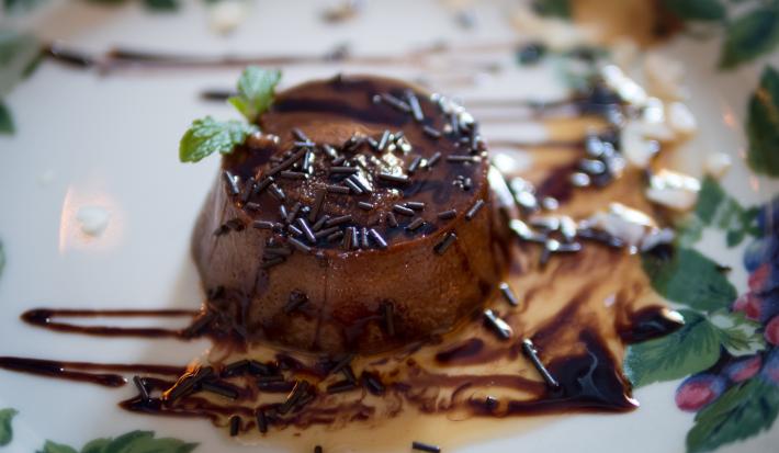 Fotografia de um mini pudim de chocolate sem furo no meio, decorado com granulado, calda de chocolate e uma folha de hortelã, sobre um prato branco desenhado..