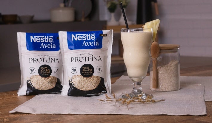 Foto da receita de shake pré-treino proteico servida em um copo de vidro alto sobre uma mesa de madeira com um pote de vidro cheio de aveia ao fundo