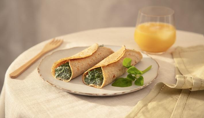 Foto em tons de bege da receita de panqueca de espinafre com ricota servida em duas porções sobre um prato de cerâmica claro em cima de uma mesa decorada com uma toalha bege, um copo de vidro com suco de laranja, um paninho verde e um garfo de madeira