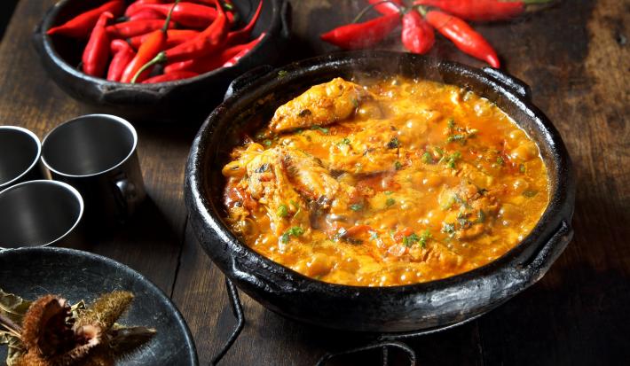Fotografia de uma panela de barro com caldeirada borbulhando de peixe com cação, postas de pescada e corvina, molho de tomate, açafrão, louro e salsinha.