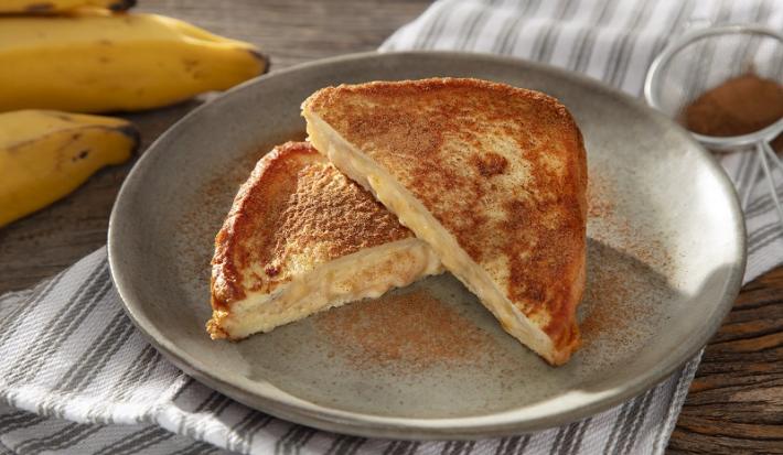 Foto em tons de cinza da receita de banana toast servida em duas metades sobre um prato de cerâmica cinza com canela em pó salpicada. Ao fundo há algumas bananas e um paninho listrado cinza
