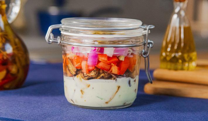 Fotografia em tons de azul com um pote de vidro ao centro. Dentro do pote existe uma salada de berinjela assada com molho de iogurte, tomate e cebola roxa