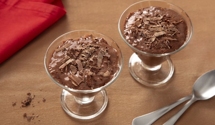 Fotografia de duas tacinhas de vidro ao lado de duas colheres de sopa. Dentro das tacinhas tem a mousse de nescau extra cacau com raspas de chocolate. Fotografia de duas tacinhas de vidro ao lado de duas colheres de sopa. Dentro das tacinhas tem a mousse de nescau extra cacau com raspas de chocolate.