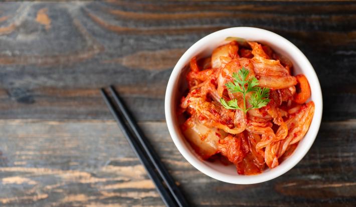 Fotografia vista de cima de um recipiente branco pequeno e fundo com uma receita de kimchi. Por cima da receita tem folhas de salsinha, e ao lado do recipiente tem dois hashis pretos sobre uma mesa de madeira escura.