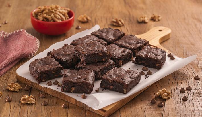 Fotografia em tons de marrom e vermelho de uma bancada de madeira com uma tábua e um papel manteiga, sobre ele pedaços de brownie. Ao fundo um recipiente vermelho pequeno com nozes e um pacote de brownie dois frades.