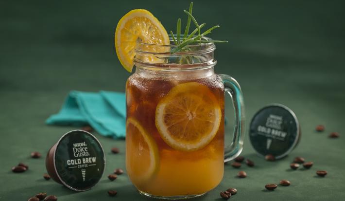 Fotografia em tons de verde de uma bancada verde, sobre ela um copo de vidro com cold brew com laranjas. Ao lado capsulas de Dolce Gusto Cold Brew e um paninho azul.