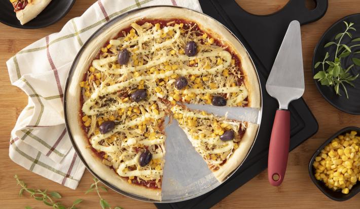 Imagem vista de cima de uma pizza redonda de frango com catupiry, milho e azeitonas em uma forma de alumínio.