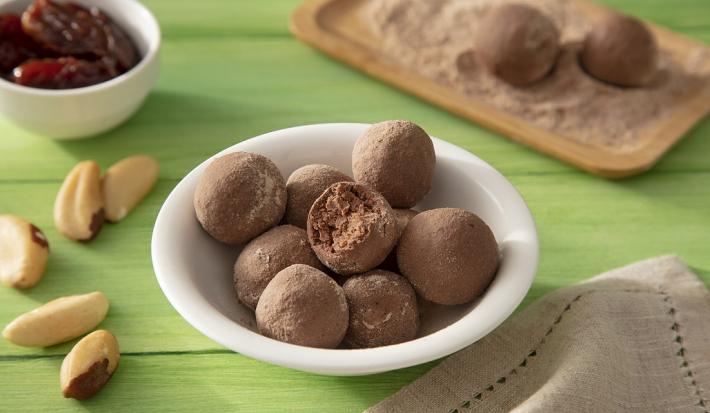 Fotografia em tons de marrom em uma bancada de madeira de cor verde. Ao centro, um bowl branco contendo as trufas. Ao lado, há algumas castanhas do pará e um pano marrom. Ao fundo, um pote com tâmaras e uma tábua com mais trufas.