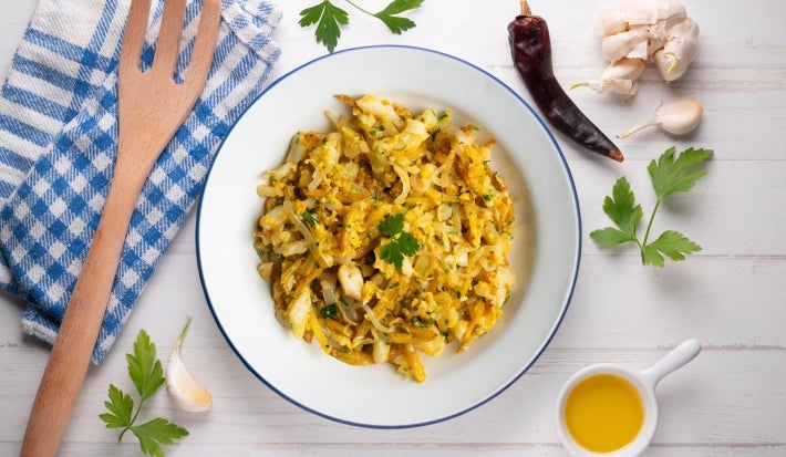 Em cima de uma mesa, prato redondo com porção de bacalhau desfiado com cebola, decorado com folha de salsa. Em torno do prato, uma colher de pau, pimenta, dentes de alho e azeite.