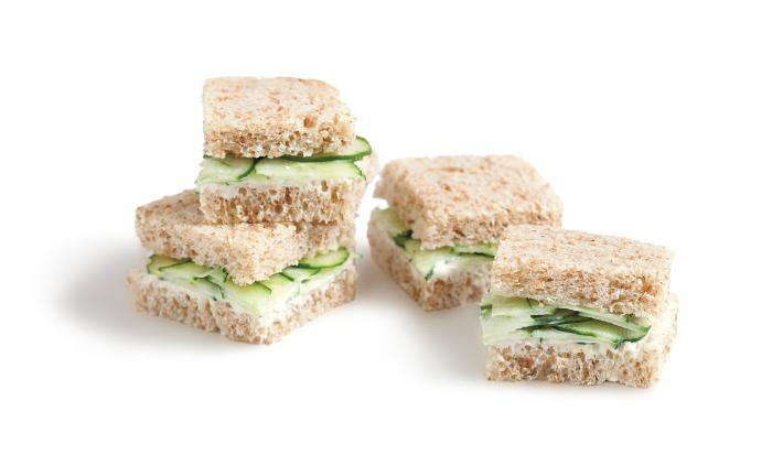Fotografia em tons de verde em um fundo branco com quatro mini sanduichinhos no pão integral com recheio de pepino.