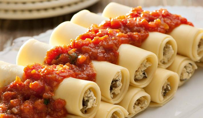 Fotografia em tons de vermelho em uma bancada de madeira com um recipiente oval branco grande com vários canelones de berinjela e com molho de tomate em cima dele.