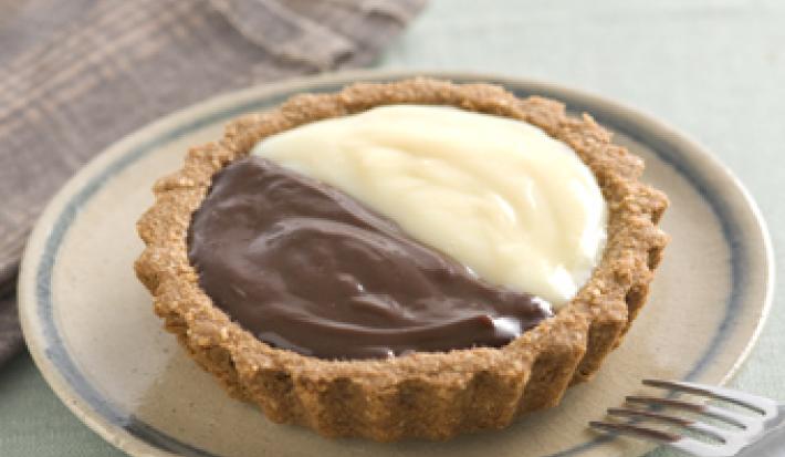 Fotografia em tons de bege em uma mesa de madeira com um pano bege xadrez, um prato redondo raso bege com detalhe azul e a tortinha de leite condensado Moça e chocolate em cima dele.