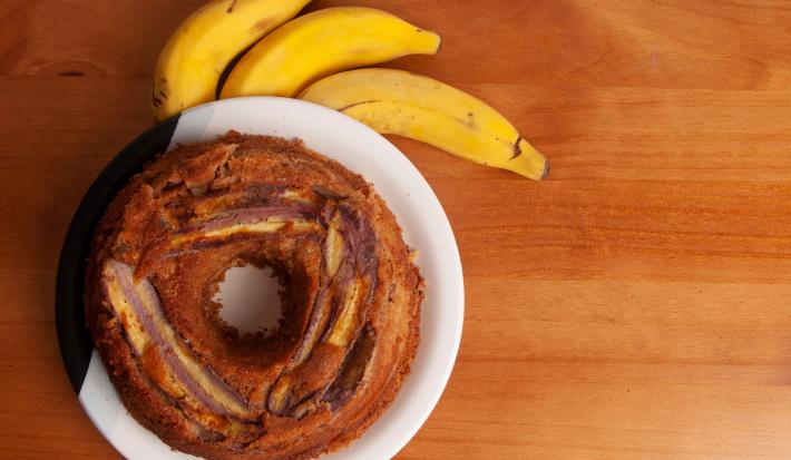 Fotografia em tons de marrom claro com um prato branco ao centro. Dentro do prato existe um bolo de banana redondo com um furo no meio, por cima do bolo existe pedaços de bananas caramelizadas. Ao lado existe três bananas inteiras.