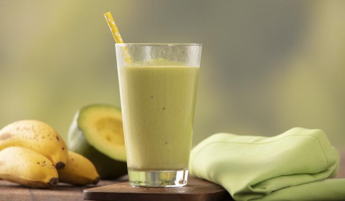 Fotografia em tons de verde e amarelo de uma bancada de madeira com um paninho verde e um apoio de copo de madeira, sobre ele um copo de vidro com a vitamina de abacate e um canudo amarelo. Ao fundo três bananas e meio abacate.