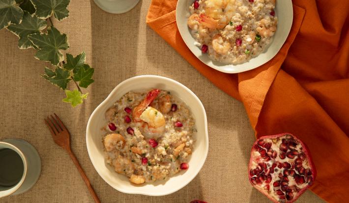 Foto da receita de risoto de camarão com brie e romã servida em um bowl de cerâmica branca sobre um paninho laranja e outro sobre uma mesa decorada com uma toalha de linho
