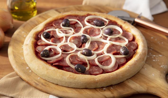 Imagem de uma pizza redonda de calabresa com cebola e azeitonas sobre uma tábua de madeira.