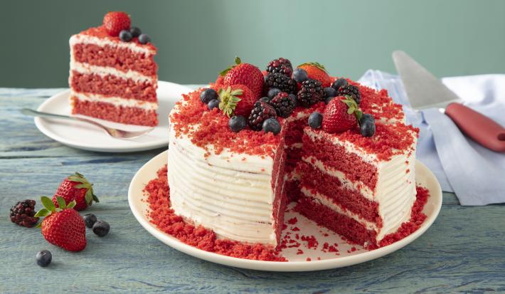 Fotografia em tons de vermelho em uma bancada de madeira clara, um prato redondo branco com o bolo red velvet cortado ao meio. Ao fundo, um prato pequeno com o pedaço.