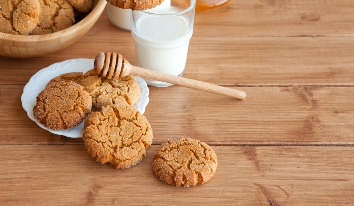 Em cima de uma mesa, prato branco com biscoitos crocantes, decorados com granulado de açúcar cristal. Ao lado do prato, copo com mel.