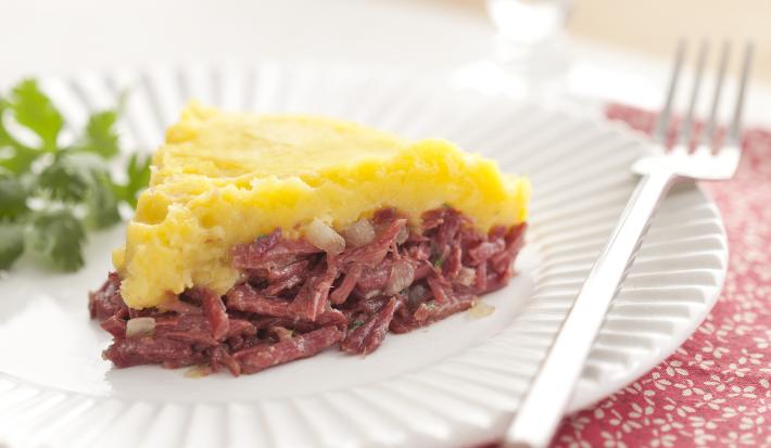 Fotografia em tons de vermelho em uma mesa de madeira clara com um prato redondo raso branco ao centro com uma fatia do escondidinho de carne seca com mandioquinha e coentro ao lado para enfeitar o prato.