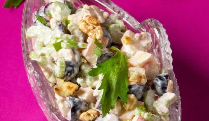 Fotografia de uma salada fresca e colorida em um prato de vidro transparente, composta por melão picado, uvas pretas, nozes e maçãs em um molho cremoso, decorada com salsa fresca, destacando a frescura.
