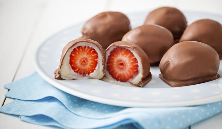 Fotografia em tons de branco de uma bancada branca, com um pano dobrado azul com bolinhas brancas, sobre ele um prato branco redondo com um bombom partido ao meio, mostrando o creme branco e o morango e cinco bombons inteiros.