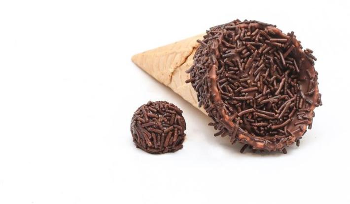 Fotografia de um cone trufado com brigadeiro sobre uma mesa off white. Ao lado do cone tem um brigadeiro passado no granulado.