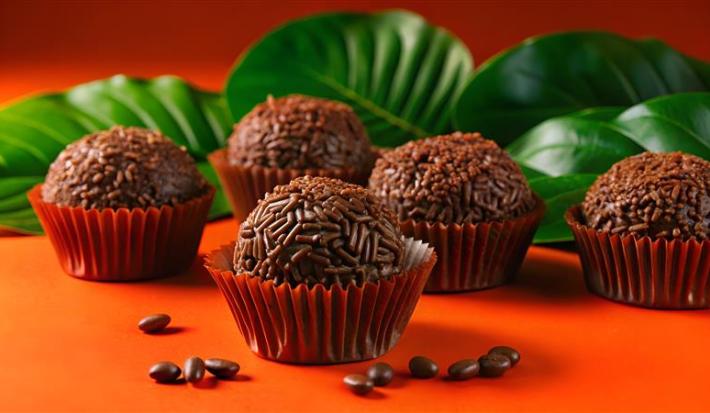 Fotografia de cinco brigadeiros passados no granulado, dentro de uma forminha vermelha, sobre uma mesa laranja com grão de café. No fundo tem algumas folhas.