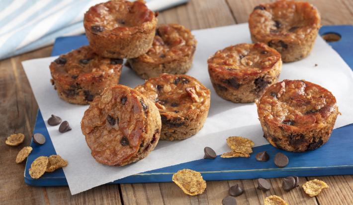 Fotografia em tons de azul em uma bancada de madeira com uma tábua azul, com um folha de papel manteiga e vários muffins de banana com gotas de chocolate em cima dela. Nesfit Cereais Matinais e algumas gotas de chocolate espalhadas pela bancada.