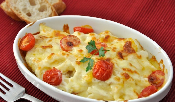 Em cima de uma mesa, refratário com batatas, queijo gratinado e tomates, decorado com folha de salsa.