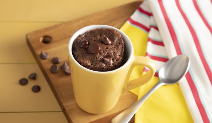 Fotografia em tons de amarelo em uma bancada de madeira amarela, um pano listrado vermelho e amarelo e uma tábua de madeira ao centro da foto com uma caneca amarela com o bolo de caneca de chocolate apoiada sobre ela.