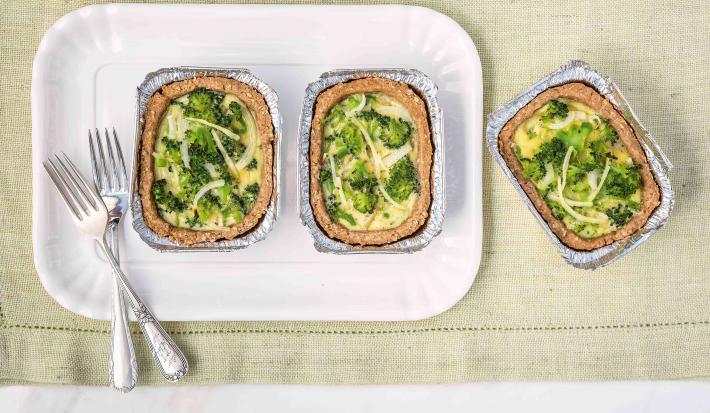 Fotografia em tons de branco, verde e dourado de uma bancada vista de cima mostrando refratário branco com duas porções de quiche com brócolis e dois garfos, com mais uma porção ao lado, tudo sobre guardanapo em tom de branco.