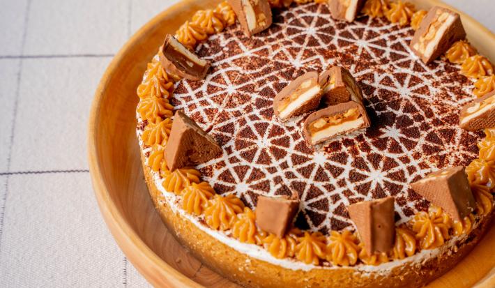 Fotografia em tons de caramelo e branco um um prato redondo de madeira ao centro. Em cima do prato existe uma torta em tons de caramelo e chocolate com alguns pedaços de chocolate no topo