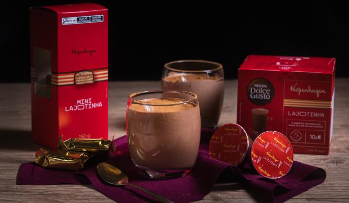 Fotografia em tons escuros de dois copos de vidro com uma receita de Mousse Cappuccino Lajotinha com cacau em pó por cima sobre um pano vinho escuro com duas cápsualsa de kopenhagen, uma colher pequena e duas lajotinhas.